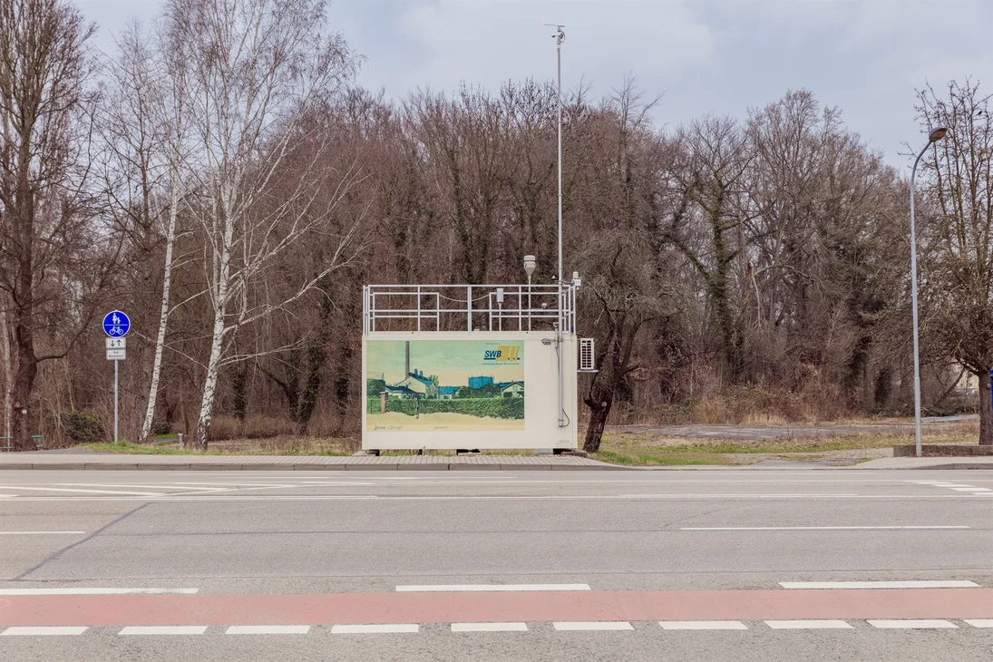 Die verkehrsnahe Messstation in Borna (weißer Container mit Probenahmeeinrichtungen auf dem Dach) steht am Rand einer Straße hinter dem Gehweg und vor Bäumen.