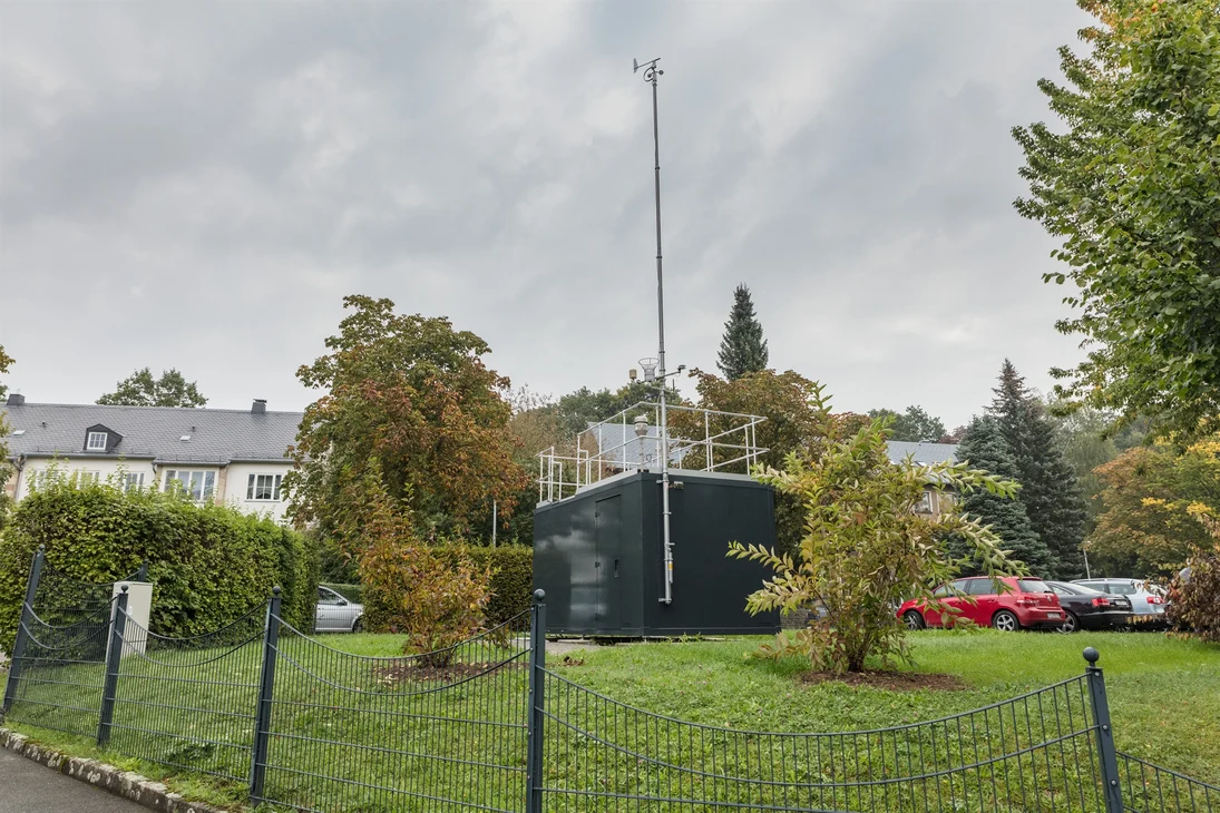 Die Messstation in der Hans-Link-Straße in Chemnitz (grüner Container mit Probenahmeeinrichtungen auf dem Dach) steht auf einer Wiese mit angrenzendem Parkplatz. Im Hintergrund sind Gebäude sichtbar.