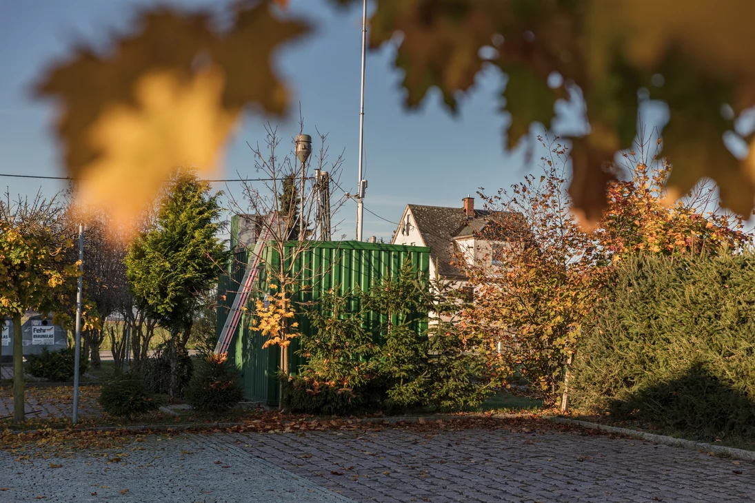 An der Messstation (grüner Container mit Probenahmeeinrichtungen auf dem Dach) lehnt eine Leiter, damit das Dach bestiegen werden kann. Neben der Messstation befinden sich Parkplätze, kleine Bäume und Glassammelsontainer. Im Hintergrund sieht man ein Haus