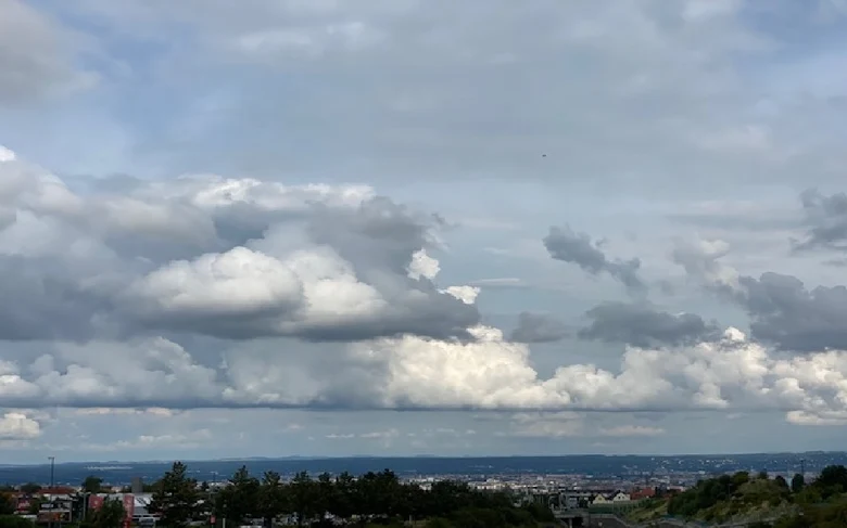 Himmel mit unterschiedlichen grauen und weißen Wolken, am unteren Bildrand sieht man Bäume, Häuser, Siedlung, Landschaft.