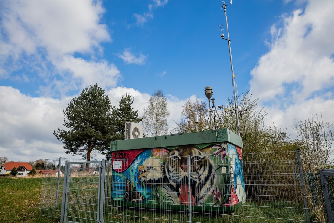 Die Messstation in Niesky (Container mit Probenahmeeinrichtungen auf dem Dach) steht auf einer Wiese. Sie ist mit Tieren bunt bemalt. Im Vordergrund dieser Malerei ist ein Tigerkopf zu sehen.