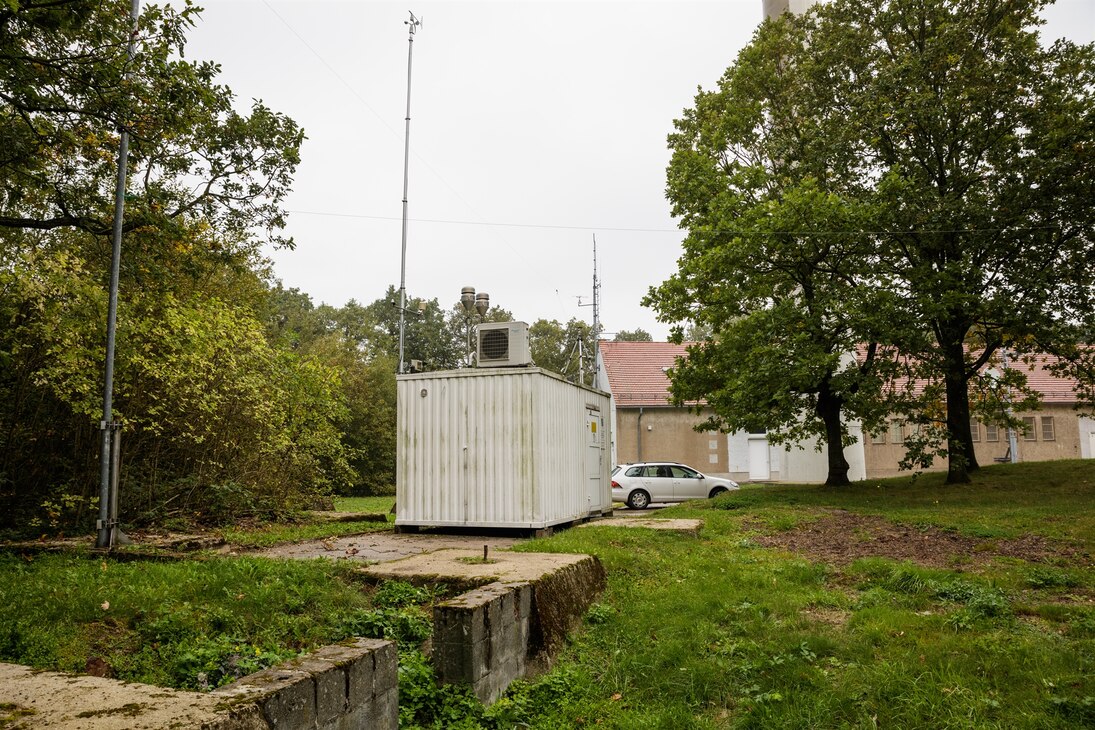 Die Messstation auf dem Collmberg (weißer Container mit Probenahmeeinrichtungen auf dem Dach) ist umgeben von grünen Wiesen, Bäumen und einem Gebäude.n